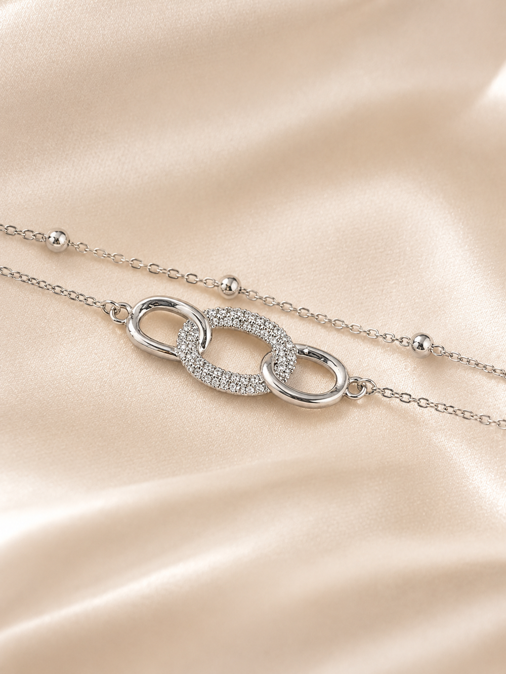 A silver-toned double-strand chain necklace with a central interlocking oval link design, one half of which is paved with small clear stones. The necklace is displayed on a soft, draped champagne-colored satin fabric. The Linked Grace Bracelet by Ashaarya, an anti-tarnish silver bracelet crafted in hypoallergenic 316L stainless steel with sparkling cubic zirconia links for lightweight everyday comfort.