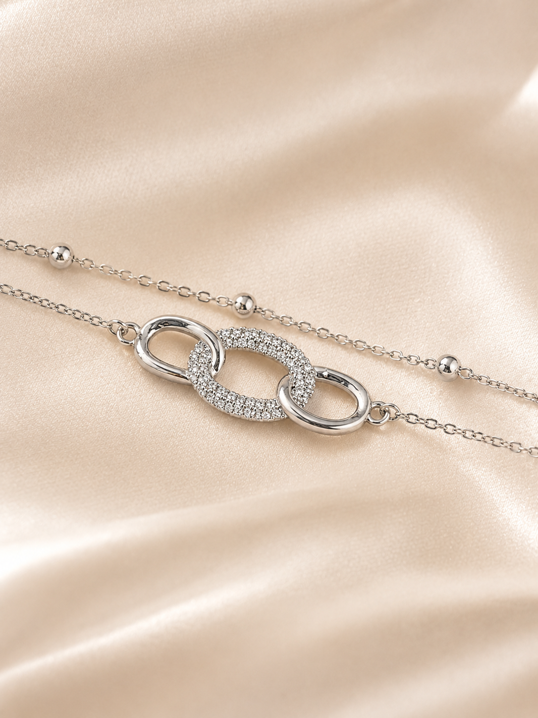A silver-toned double-strand chain necklace with a central interlocking oval link design, one half of which is paved with small clear stones. The necklace is displayed on a soft, draped champagne-colored satin fabric. The Linked Grace Bracelet by Ashaarya, an anti-tarnish silver bracelet crafted in hypoallergenic 316L stainless steel with sparkling cubic zirconia links for lightweight everyday comfort.