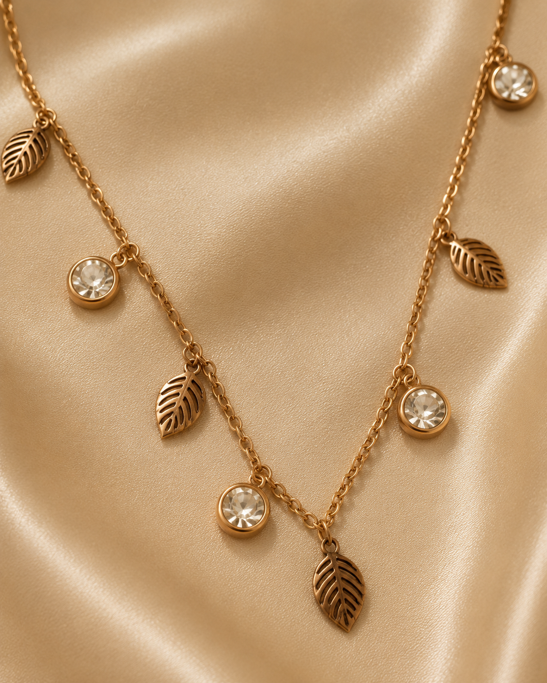 A gold-toned necklace with leaf-shaped charms and round clear crystal pendants, displayed on a draped beige satin fabric. rose gold necklace for women, anti tarnish necklace, leaf pendant necklace, nature inspired necklace, lightweight necklace for women, stainless steel necklace, 18kt rose gold necklace, dainty necklace for women, everyday necklace, minimalist jewellery, comfort jewellery, waterproof necklace, skin friendly necklace, Ashaarya necklacebackground.