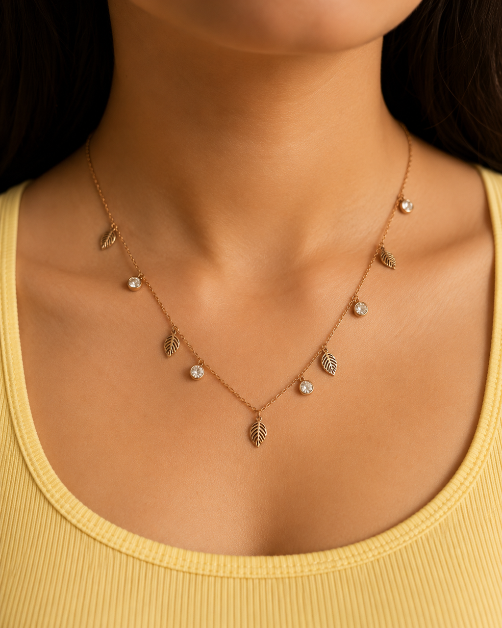 A close-up of a model wearing a delicate rose gold necklace with small leaf charms and clear rhinestone accents, paired with a yellow ribbed tank top. rose gold necklace for women, anti tarnish necklace, leaf pendant necklace, nature inspired necklace, lightweight necklace for women, stainless steel necklace, 18kt rose gold necklace, dainty necklace for women, everyday necklace, minimalist jewellery, comfort jewellery, waterproof necklace, skin friendly necklace, Ashaarya necklace