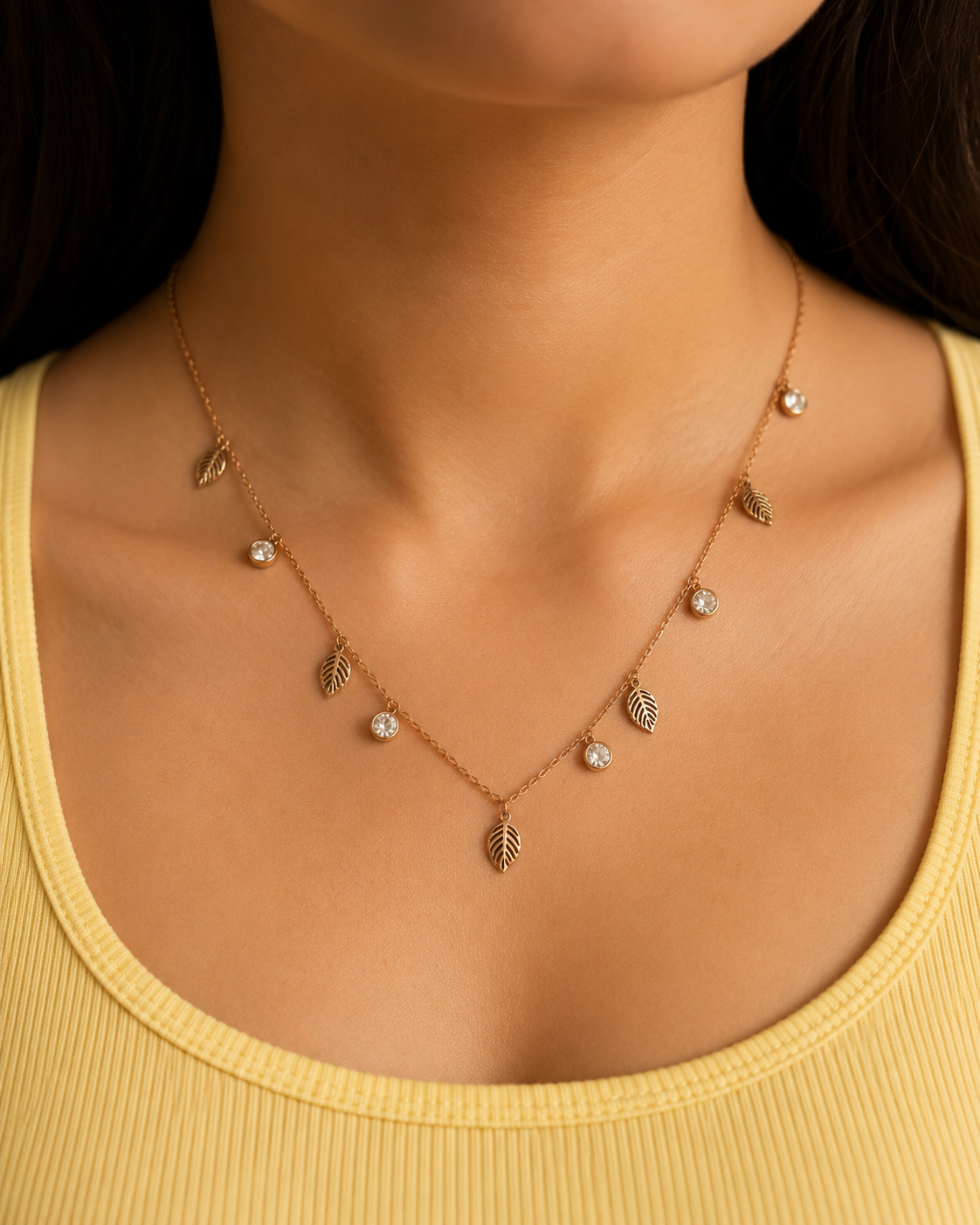 A close-up of a model wearing a delicate rose gold necklace with small leaf charms and clear rhinestone accents, paired with a yellow ribbed tank top. rose gold necklace for women, anti tarnish necklace, leaf pendant necklace, nature inspired necklace, lightweight necklace for women, stainless steel necklace, 18kt rose gold necklace, dainty necklace for women, everyday necklace, minimalist jewellery, comfort jewellery, waterproof necklace, skin friendly necklace, Ashaarya necklace