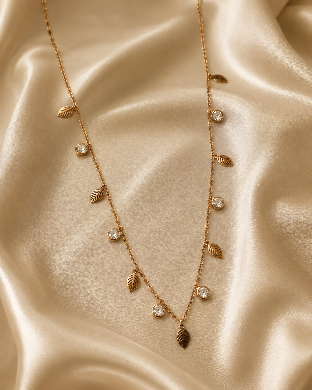 A gold leaf necklace with clear crystal accents, draped elegantly on a shimmering beige satin fabric background. rose gold necklace for women, anti tarnish necklace, leaf pendant necklace, nature inspired necklace, lightweight necklace for women, stainless steel necklace, 18kt rose gold necklace, dainty necklace for women, everyday necklace, minimalist jewellery, comfort jewellery, waterproof necklace, skin friendly necklace, Ashaarya necklace